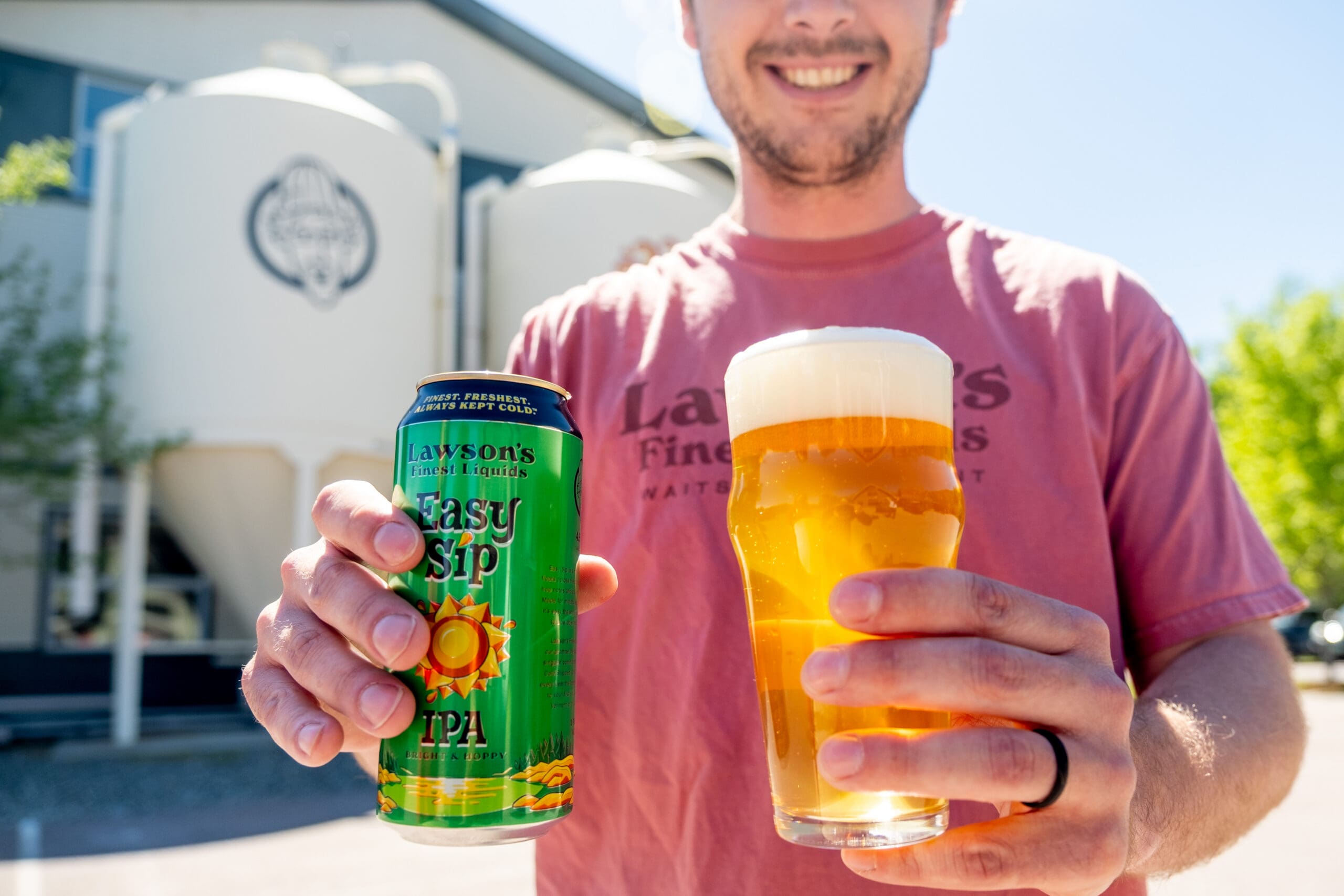 Man smiling, holding a can of Lawson’s Finest Liquids Easy Sip IPA and a glass of beer, with brewery tanks in the background under a clear blue sky.