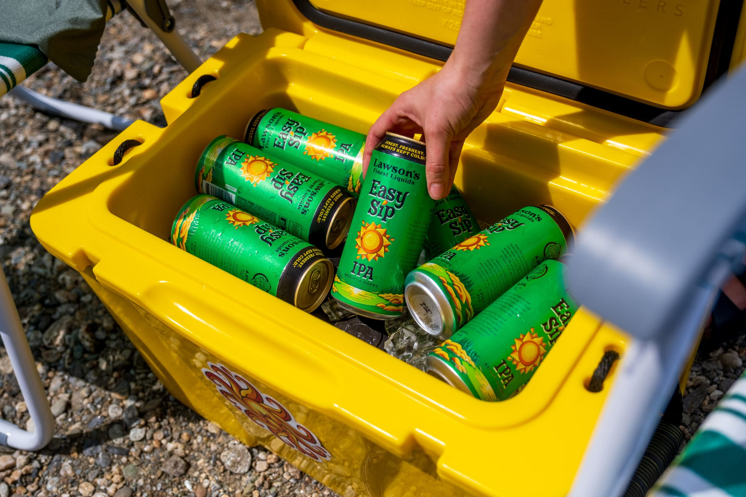 A hand reaches into a yellow cooler filled with green cans of Easy Sip IPA. The scene depicts a casual outdoor gathering on a sunny day.