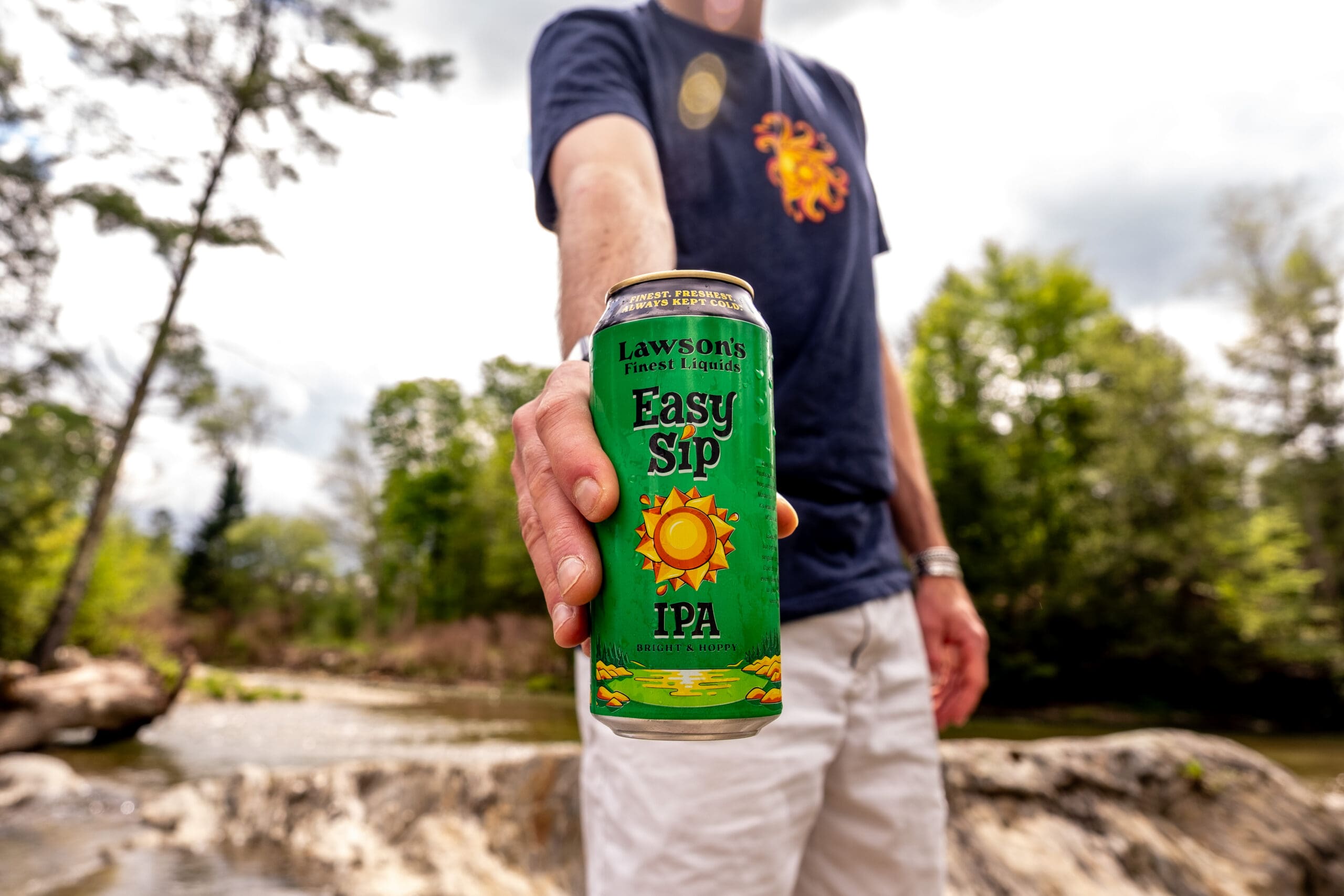 A person in a navy shirt and white shorts holds a can of Lawson's Finest Liquids Easy Sip IPA towards the camera. They're outdoors near a river with trees in the background, creating a relaxed, nature-focused vibe.