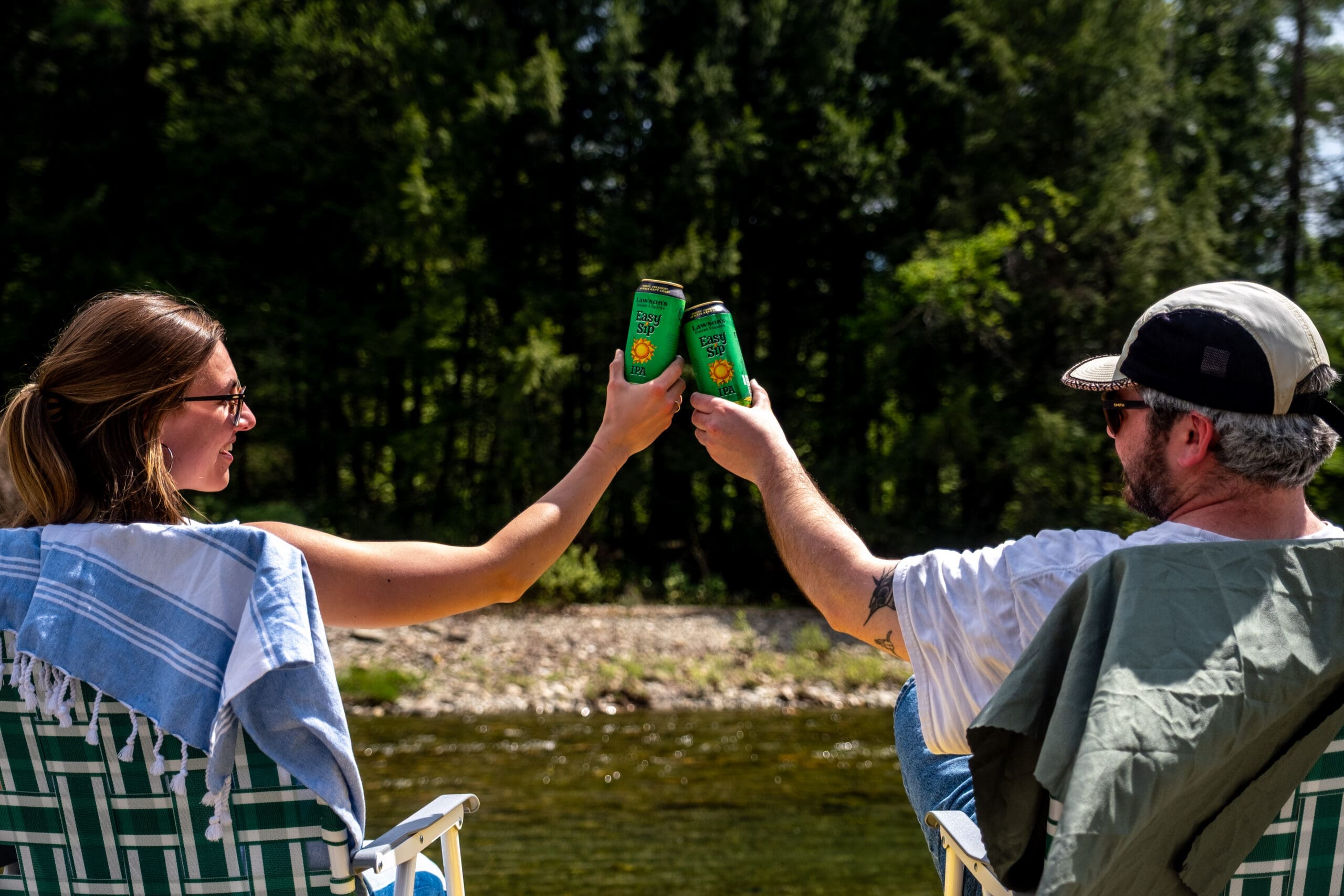 A woman and a man sit in chairs by a riverside, toasting with green cans of Easy Sip IPA. The mood is relaxed and joyful, with a forest in the background.