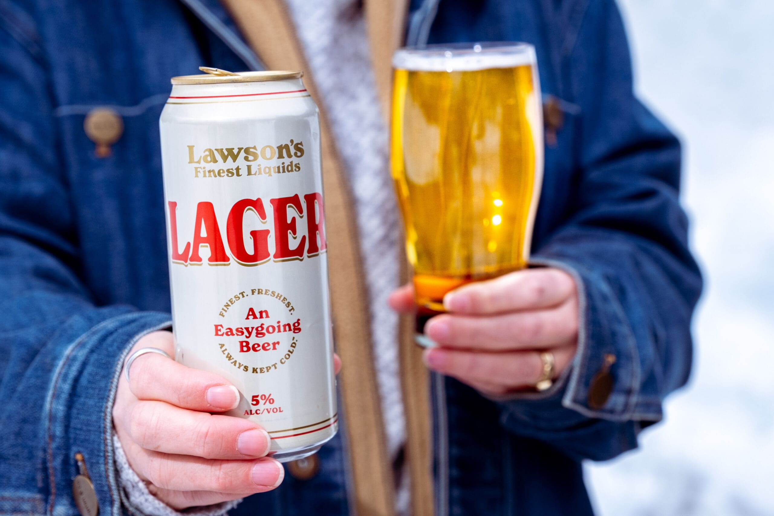 A person in a denim jacket holds a can of Lawson's Finest Liquids Lager and a glass filled with golden beer. The label reads “Lawson's Finest Liquids, Lager, An Easygoing Beer.”
