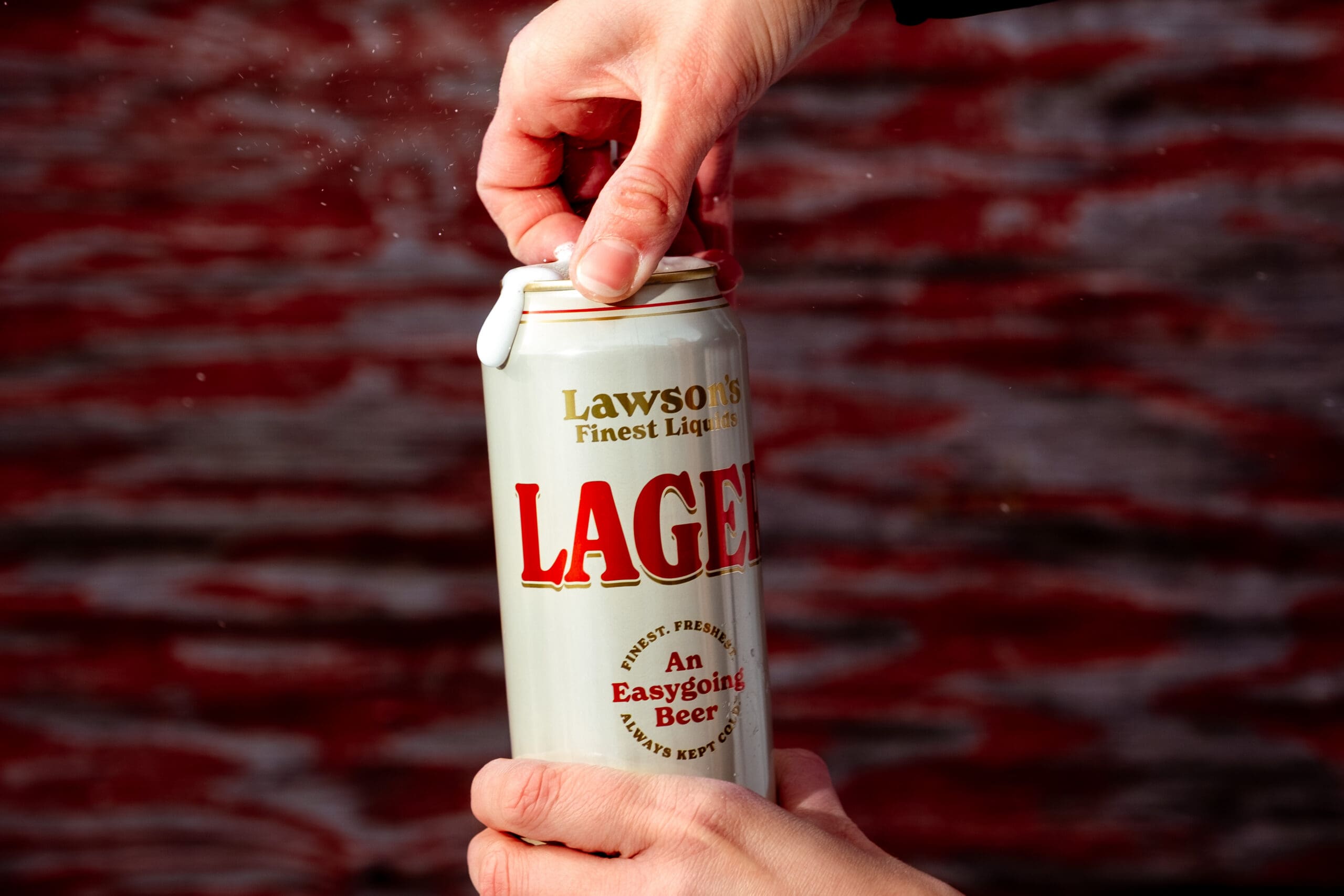 Hands opening a tall can of Lawson's Finest Liquids Lager, with foam emerging. The background is a blurred, reddish-brown texture, conveying a casual tone.