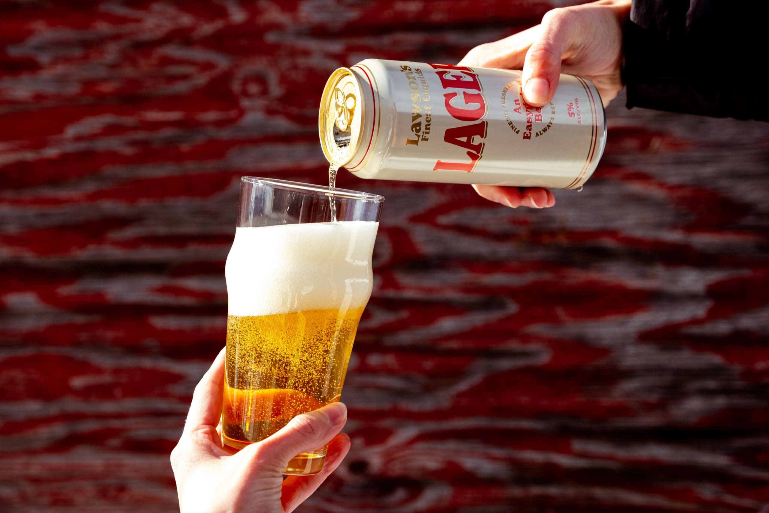 Person pouring a Lawson's Finest Liquids Lager from a can into a pint glass, forming a frothy head. Background is a textured wooden surface in red and brown.