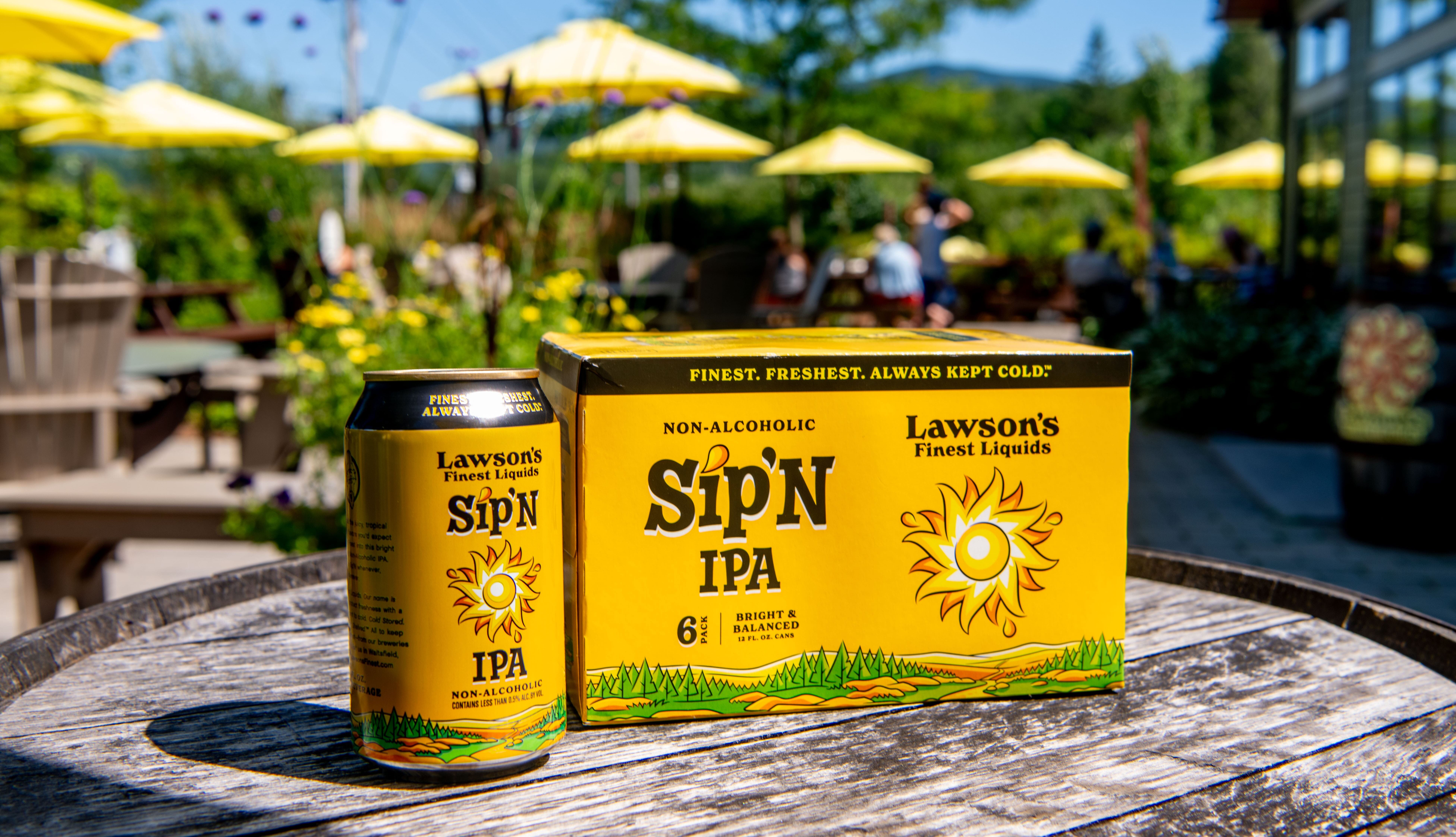 Lawson’s Finest Liquids Sip'N IPA non-alcoholic beer can and box on a sunny patio table, with yellow umbrellas and greenery behind.