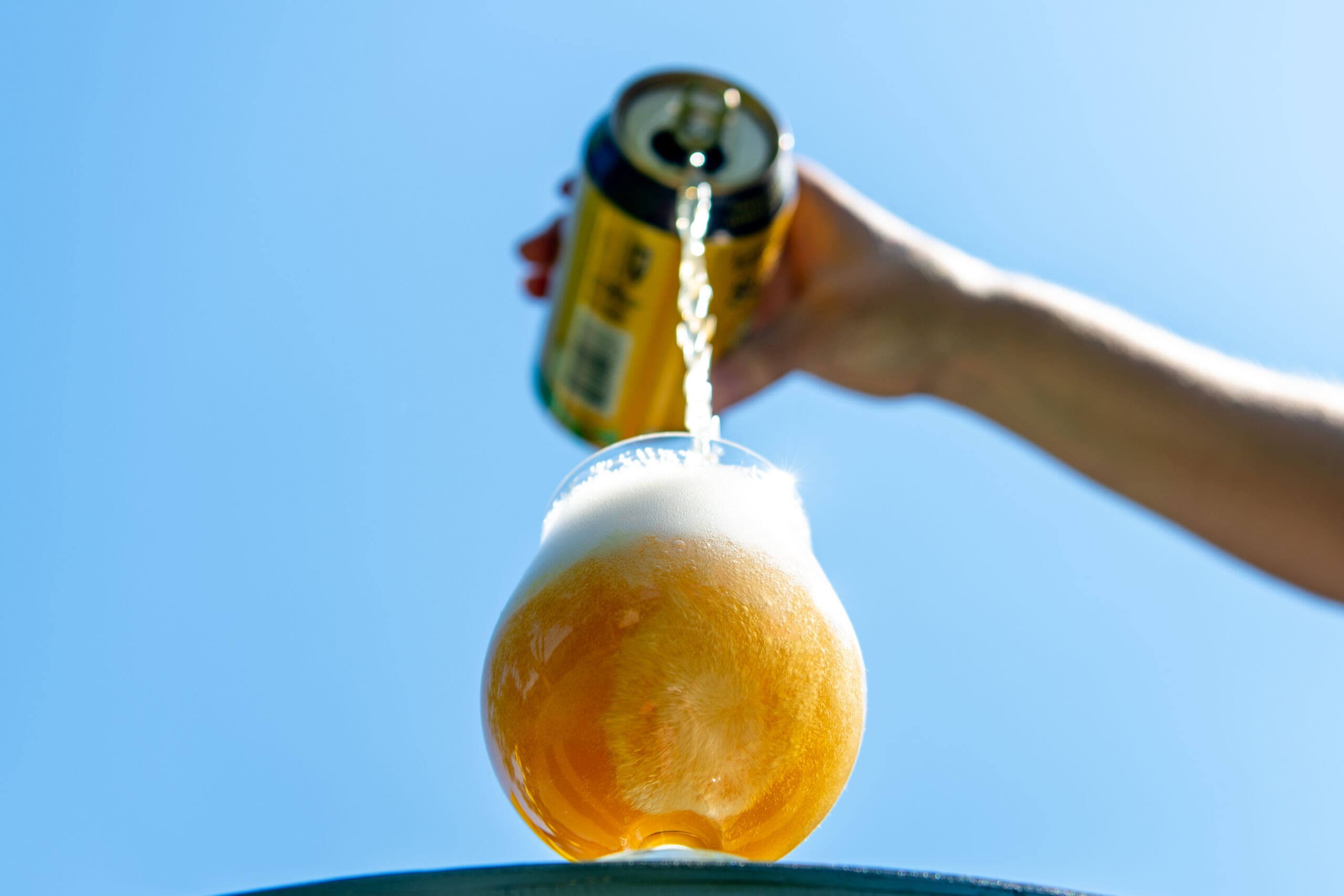A hand pours golden beer from a can into a rounded glass, creating a foamy head against a bright blue sky in a refreshing outdoor scene.