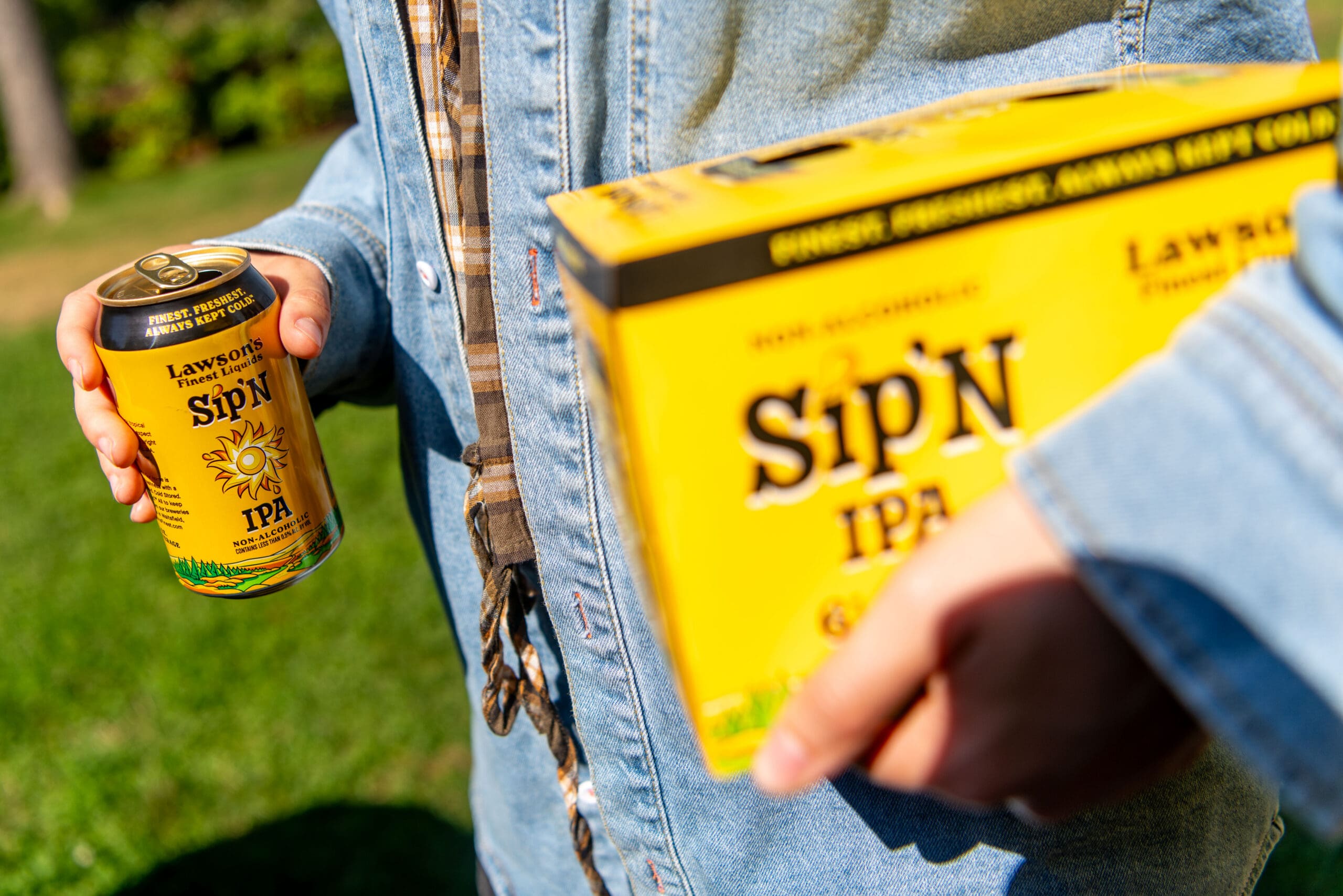 Person in a denim jacket holds a can and six-pack box of Lawson’s Finest Liquids Sip'N non-alcoholic IPA outdoors, suggesting a sunny, casual outing.