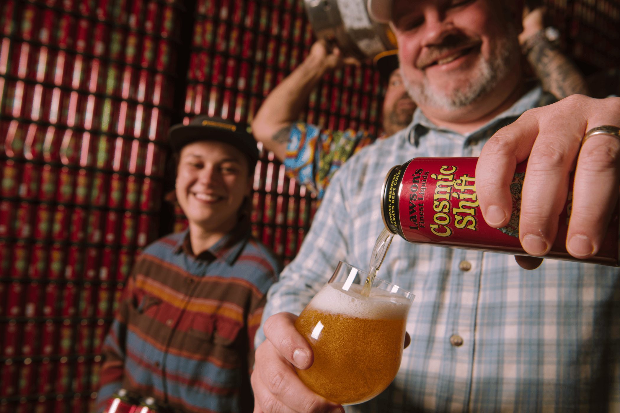 A smiling man pours Cosmic Shift IPA from a red can into a glass, with a woman and colorful rows of cans behind him in a lively brewery warehouse.